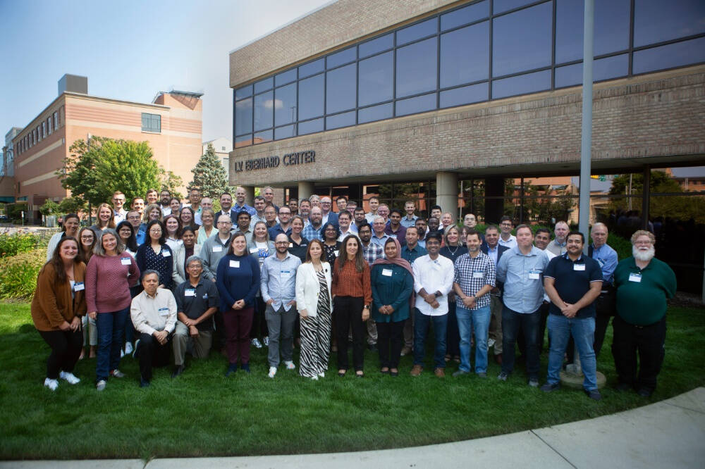 Faculty and staff stand together on a green lawn in front of the  WM EBERHARD CENTER. The group is arranged in several rows, with some people standing and a few seated in front.
